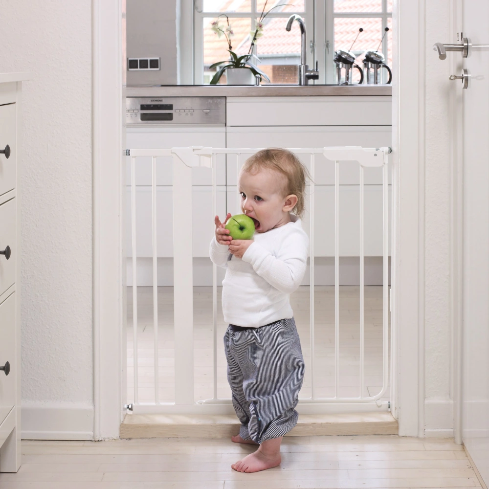 Toddler stands at a white safety gate in a doorway while eating a green apple
