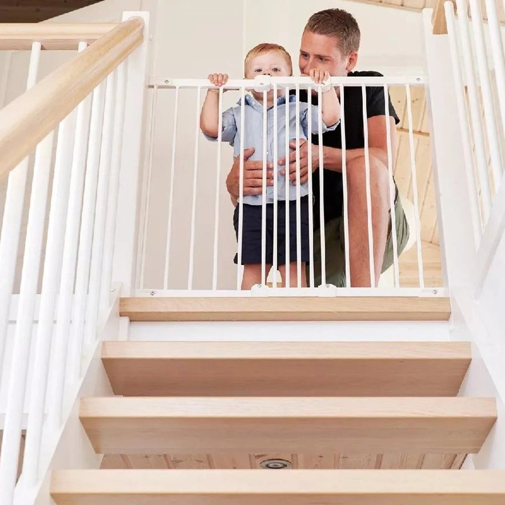 Man kneels behind a toddler at the top of wooden stairs while holding a white safety gate